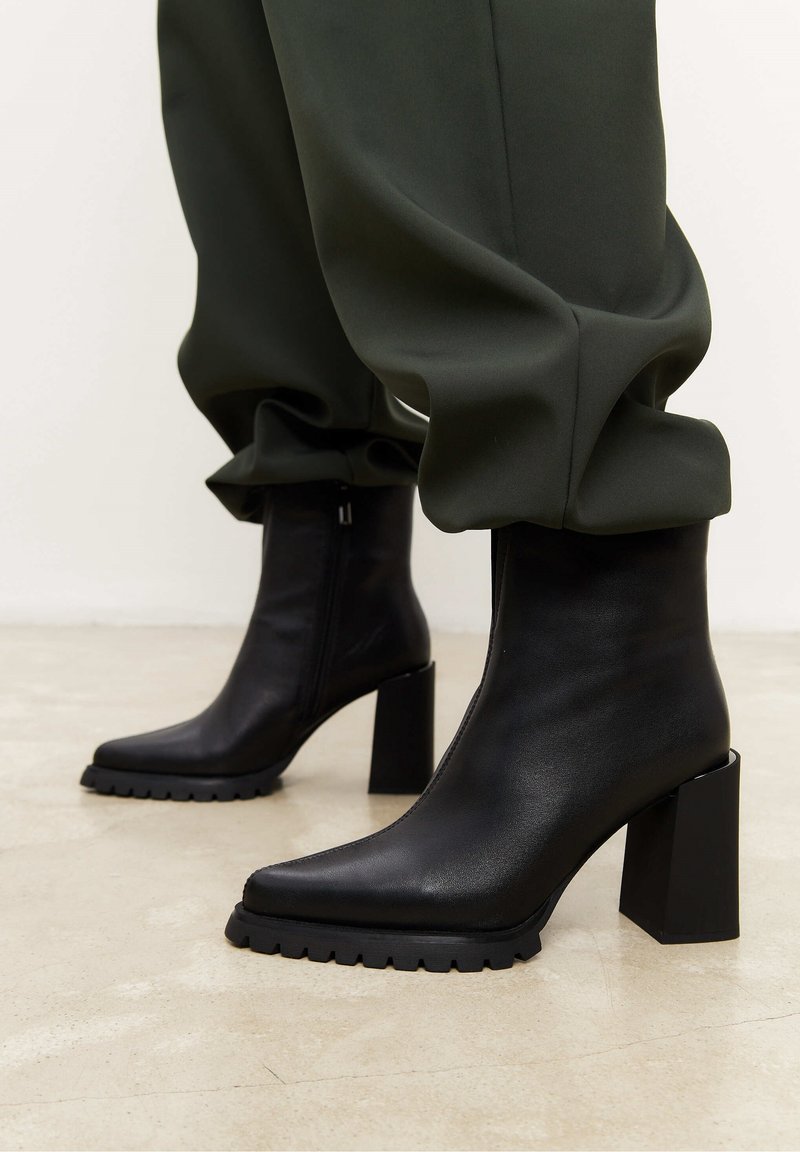 Bottes basses en cuir noir avec talons épais et semelles crantées portées avec un pantalon vert foncé retroussé sur un sol clair.
