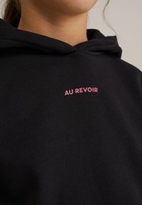 Črn bombažni pulover s kapuco z mehkimi vlakni, ki na prednjem delu prikazuje rožnato vezeno besedilo "AU REVOIR". Enostaven dizajn, nastavljiva kapuca.