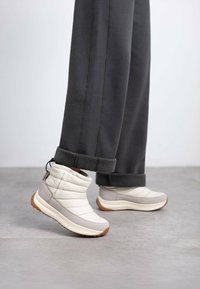 Botas acolchadas blancas y gris claro hasta el tobillo con un diseño texturizado, que cuentan con una suela de goma y una pestaña en la parte trasera, combinadas con pantalones grises.