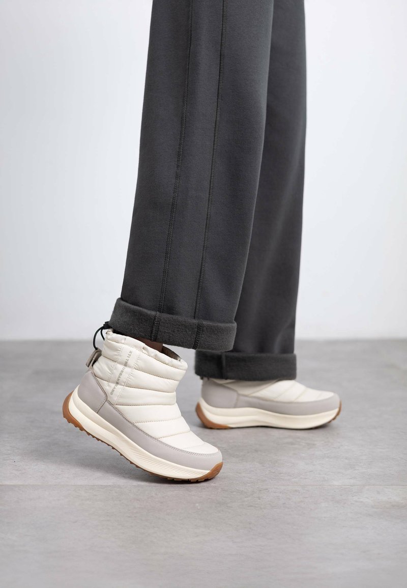 Botas acolchadas blancas y gris claro hasta el tobillo con un diseño texturizado, que cuentan con una suela de goma y una pestaña en la parte trasera, combinadas con pantalones grises.