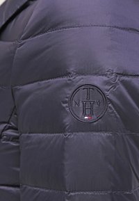 Mörkblå quiltad jacka med rund broderad logotyp som har bokstäverna T, N, H, Y och en liten röd, vit och blå flagga.