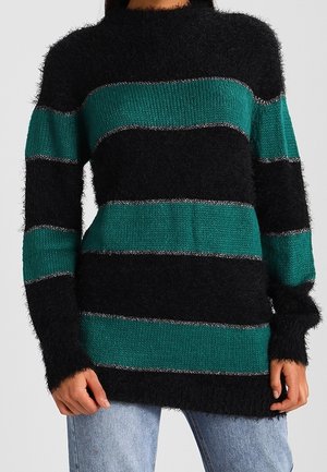 Tynd, sort og grønstribet langærmet sweater brugt sammen med lyseblå jeans, vist fra halsen til hofterne.