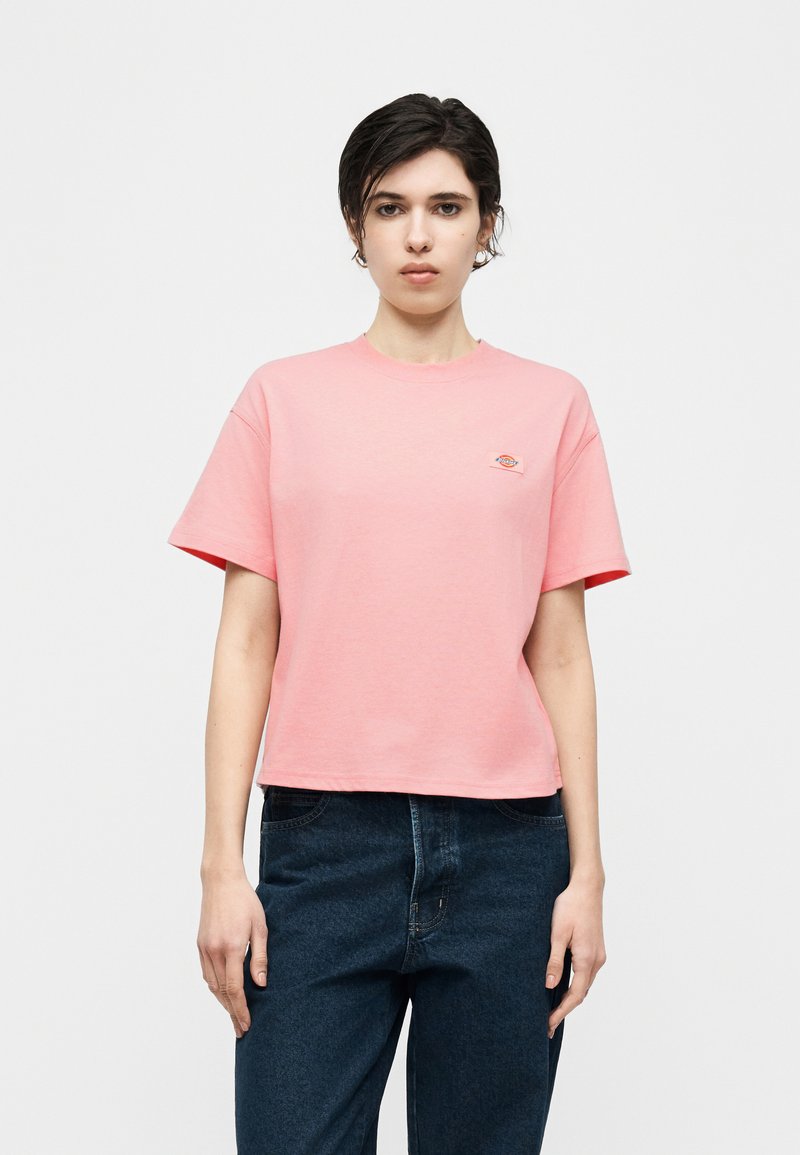 Jonge persoon met kort donker haar, gekleed in een roze T-shirt met korte mouwen en donkerblauwe spijkerbroek, staand tegen een effen witte achtergrond.