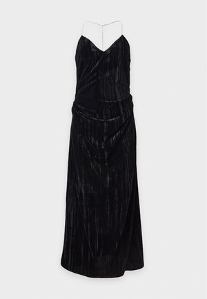 Une femme porte une longue robe noire avec un sac à main à motifs et des ballerines noires pointues, sur un fond blanc uni.