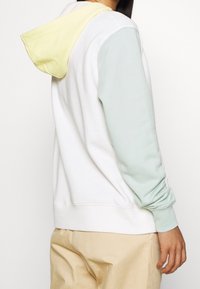 Bílá mikina s kapucí s pastelově zelenými a žlutými akcenty na rukávech a kapuci. Měkká látka a volný střih s žebrovanými manžetami.