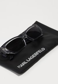Lunettes de soleil noires avec une monture transparente reposant sur une pochette noire avec le logo blanc "KARL LAGERFELD". Design angulaire avec des verres foncés.