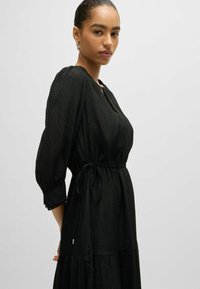 Schwarzes, strukturiertes Kleid mit drei Viertelärmeln, elastischem Taillenzug und gestuftem Saum. Ein Schlitzdetail am Vorderteil sorgt für einen subtilen Akzent.