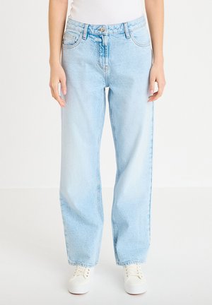 Persona che indossa jeans a gamba dritta azzurri e scarpe da ginnastica bianche, in piedi davanti a uno sfondo bianco uniforme.