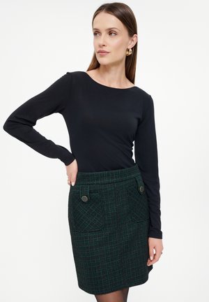 Zwart langemouwen top gecombineerd met een groen geruit minirok met zijzakken en knopen voor detail. De gladde textuur contrasteert met de wolachtige stof.