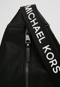 Czarna torba Michael Kors z widocznym srebrnym zamkiem błyskawicznym i szerokim czarnym paskiem z białym napisem "Michael Kors".