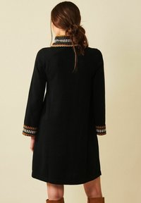 Nainen, jolla on ruskeat hiukset löysässä nutturassa, pukeutuneena mustaan pitkähihassa olevaan mekkoon, jossa on kuvioitu kaulus ja hihat, seisoo vaaleanbeigeä taustaa vasten.