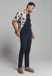 Korte mouwen bloemenoverhemd in crème met blauwe en bruine bloemen, marineblauw tanktop, donkere broek en bruine sandalen met dubbele banden.