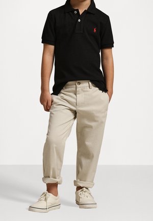 Czarna koszulka polo z czerwonym logo, beżowe spodnie z podwiniętymi nogawkami i białe sneakersy, prezentujące swobodny, dopasowany styl oraz miękkie tekstury materiału.
