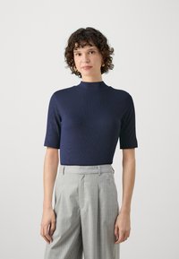 Marineblå topp med korte ermer og høy hals, med ribbestruktur. Kombinert med lysegrå pinstripe bukser som viser en skreddersydd passform.