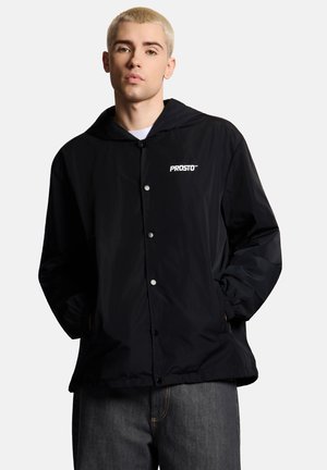 Młoda osoba z krótkimi blond włosami, ubrana w czarną kurtkę z kapturem z logo "PROSTO", ręce w kieszeniach i ciemnoszare spodnie.