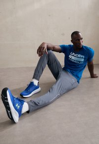 Modrá športová tričko s logom "UNDER ARMOUR", šedé jogger nohavice a modré tenisky s čiernymi podrážkami a zelenými akcentmi, na svetlom pozadí.