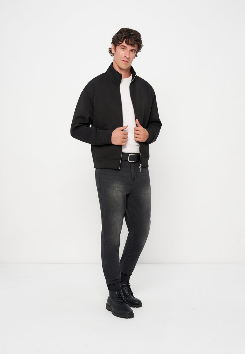 Mann, der eine schwarze Zip-up-Jacke, ein weißes Hemd, schwarze verwaschene Jeans und schwarze Stiefel trägt, steht vor einem schlichten weißen Hintergrund.