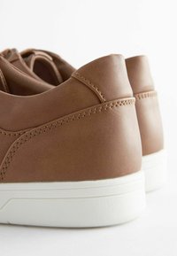Bruna lädersneakers med en slät yta, vita gummisulor och detaljerad sömnad längs sömmarna. Bakifrån framhävs en rundad häl design.
