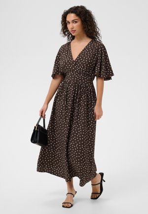 Femme aux cheveux bouclés portant une robe midi marron à pois, des sandales à talons noires et tenant un petit sac à main noir.