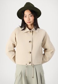 Beige cropped jasje met grote knopen, zijzakken en een overhemdkraag. Gepaard met een groene hoed en een lichtgroene rok met plooien.
