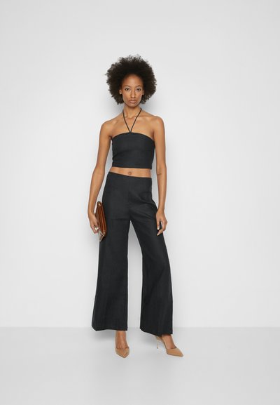 Top halter preto curto com decote reto, combinado com calças largas pretas. Modelo segurando uma carteira castanha e usando sapatos de salto bege.