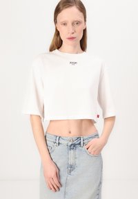 Balta trumpa medvilninė marškinėlių palaidinė su trumpomis rankovėmis ir juodu "JOOP! JEANS" logotipu ant krūtinės. Modelis vilki šviesią džinsinę sijoną.