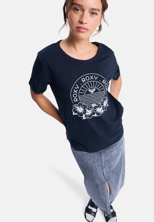 T-shirt en coton bleu marine avec un design graphique blanc représentant un soleil et des fleurs. Associé à une jupe en denim clair avec une fente à l'avant. Sandales noires.