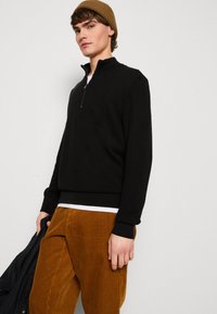 Black pullover sweater with a quarter zip, ribbed cuffs, and hem. Worn with textured brown corduroy pants. Model holding a jacket.
