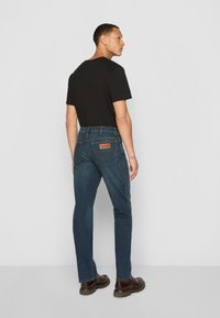 Dark denim jeans with a straight fit, five-pocket design, and a leather patch on the back. Paired with a black T-shirt and brown shoes.