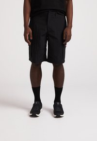 Schwarze Shorts aus strukturiertem Stoff, mit Knopfverschluss vorne und Seitentaschen. Kombiniert mit schwarzen Sportschuhen und Socken.
