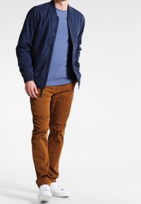 Giubbotto bomber navy sopra una t-shirt azzurra chiara, abbinato a pantaloni marroni e sneakers bianche. Presenta una chiusura con zip e polsini a costine.