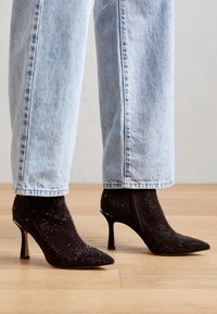 Stivaletti neri con tacco alla caviglia e punta appuntita, caratterizzati da una superficie testurizzata decorata con piccole borchie, abbinati a jeans in denim blu.