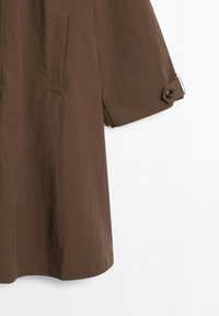 Brown knee-length coat with side slit pocket and short sleeve featuring a buttoned tab detail against white background.