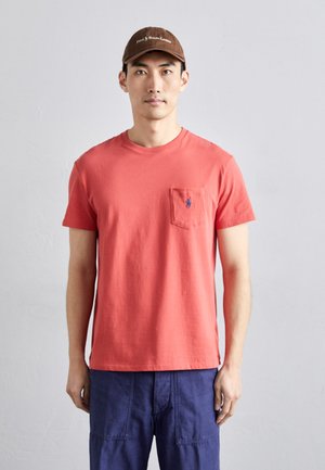 Koraalkleurig katoen T-shirt met een ronde hals en een borstzak, voorzien van een klein blauw logo. Gekleed met marineblauwe broek.