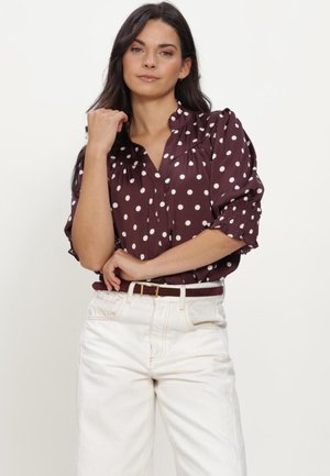 Vrouw die een donkerpaarse blouse met witte stippen en witte hoge taillebroeken draagt, poseert met één hand bij haar gezicht.