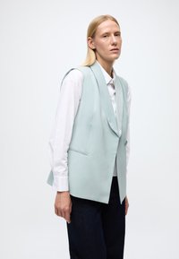 Blazer bleu deschis fără mâneci, cu rever tip șal, închidere cu un singur nasture și buzunare laterale, asortat cu o cămașă albă cu nasturi și pantaloni închiși la culoare.