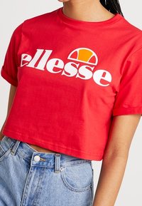 Camiseta de algodón roja recortada con letras blancas "ellesse" y un logo curvado naranja. Manga corta, cuello redondo, textura suave.