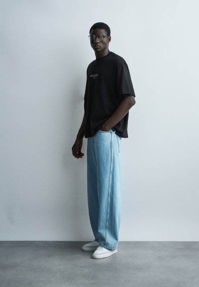 Νεαρός άνδρας με γυαλιά, μαύρο υπερμεγέθες t-shirt, ανοιχτό μπλε τζιν παντελόνι με φαρδιά πόδια και λευκά αθλητικά παπούτσια, στέκεται μπροστά σε έναν απλό ανοιχτό τοίχο.