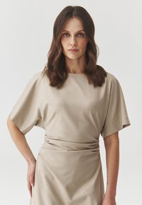 Beige mekko lyhyillä hihoilla, jossa on pyöreä kaula-aukko ja kerätty vyötärö. Sileä kangas, jossa on hieman kiiltoa, laskostava siluetti.