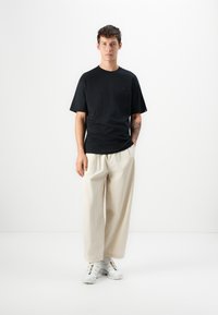T-shirt preto de manga curta, corte descontraído, combinado com calças largas em cor creme. Ténis brancos com detalhes coloridos. Fundo minimalista.