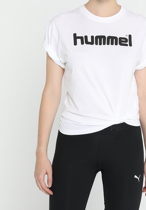 Osoba ubrana w białą koszulkę "hummel" z podwiniętymi rękawami oraz czarne legginsy Puma na jednolitym jasnym tle.