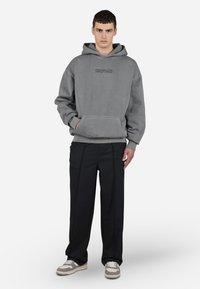 Sudadera con capucha gris con bolsillo frontal y logotipo bordado, combinada con pantalones negros y zapatillas beige. Textura de tela suave.