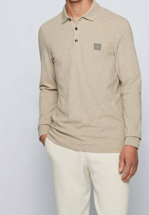 Homme portant un polo beige à manches longues avec trois boutons et un patch logo sur la poitrine, associé à un pantalon blanc cassé, debout avec une main dans la poche.