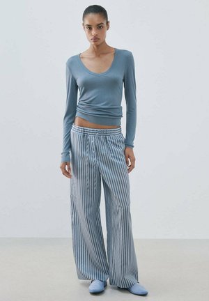 Joven mujer de pie, lleva puesta una camiseta azul ajustada de manga larga, pantalones sueltos azul y blanco con rayas verticales y zapatos azul claro sin cordones.