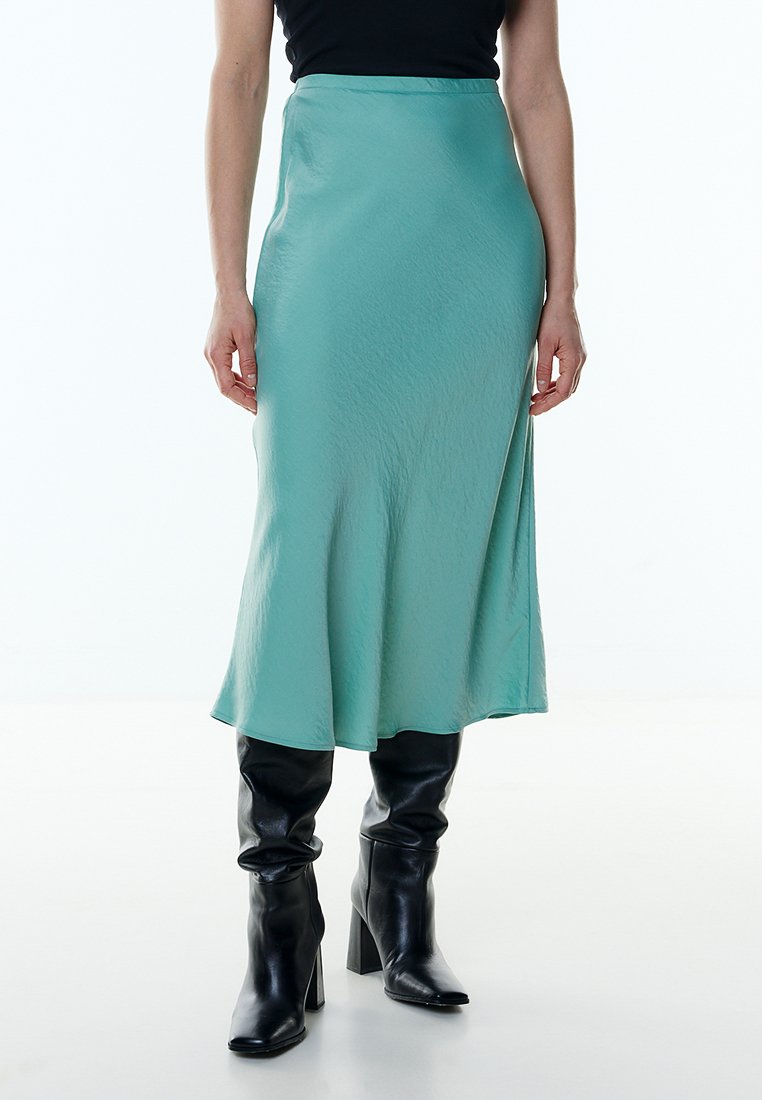 Woman wearing a mint green midi skirt paired with black heeled knee-high boots and a black sleeveless top, against a white background.
