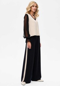 Gebreide beige top met zwarte doorschijnende mouwen, gecombineerd met zwarte wijde broek met een beige streep. Soepele textuur en relaxte pasvorm.