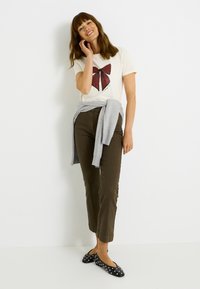 Camiseta crema con gráfico de lazo de rayas rojas, pantalones verde oliva, suéter gris atado a la cintura, bailarinas negras con lunares blancos.