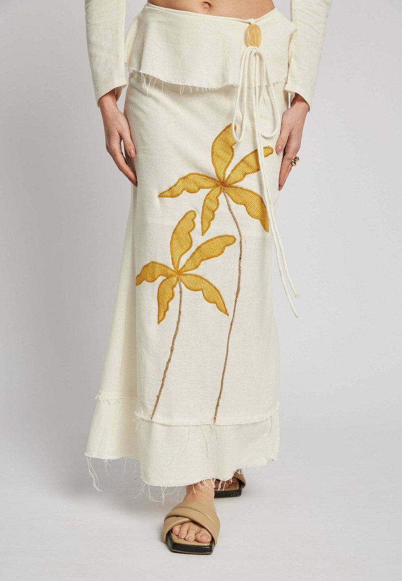 White linen maxi skirt with yellow embroidered palm leaves, frayed hem, and drawstring waist. Paired with beige slide sandals.