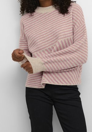 Vrouw die een roze-beige gestreepte trui draagt en de mouwomslag aanpast, gecombineerd met een zwarte broek tegen een effen witte achtergrond.