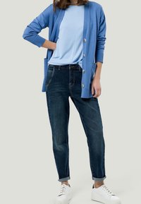 Hellblauer Cardigan mit Knöpfen, über einem passenden blauen Top getragen. Dunkelblaue Jeans mit umgeschlagenen Säumen und weiße Sneakers vervollständigen das Outfit.